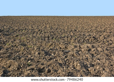 A rough stony field lies ploughed and cultivated ready for sowing spring crops, on a sunny hillside backed by a hazy blue sky.