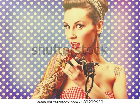 Photo of beautiful pin-up girl with tattoos, tattooing herself and looking at the camera against blue polka dot background. Retro styled imagery, grungy, toned image, noise added, vintage.
