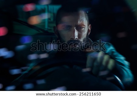 Serious young man driving a car at night. Bokeh lights reflecting from the windshield, motion blur