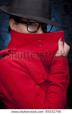 Mysterious portrait of a young woman in red trench coat, with glasses, and blue background.