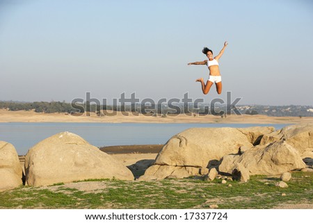 A woman jumps for joy on to of a rock overlooking a lake