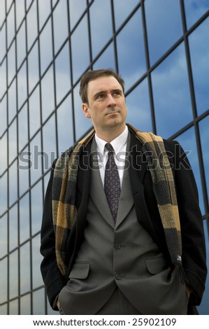 a man stands in front of a glass building wearing scarf and top coat