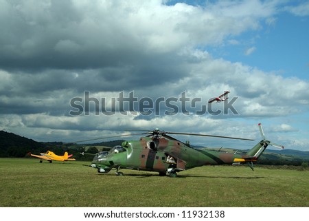 army helicopter and yellow plane on green airfield, white airplane in air, heavy clouds