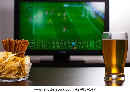 Pint of beer, chips and salty sticks on the table in front of television show of football. Set of snacks and beverage soccer fan at home.