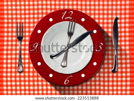 Lunch Time Concept / Clock composed by a white and red plate with fork and knife on red and white tablecloth with silver cutlery. Lunch time concept