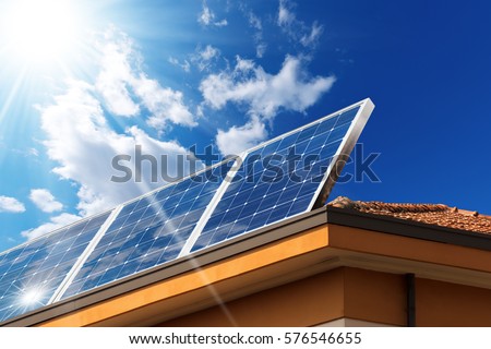 Close-up of a house roof with a solar panels on top, on a blue sky with clouds and sun rays