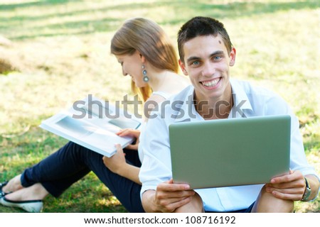 Happy young student with laptop and a girl at the background at the park