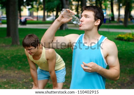 Young sportsman drinking water at the park with another person resting on the background Young sportsman drinking water at the park with another person resting on the background