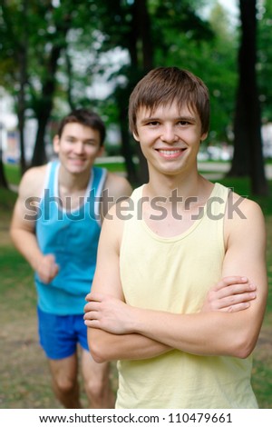 Smiling young man standing with another person jogging at the background at the park