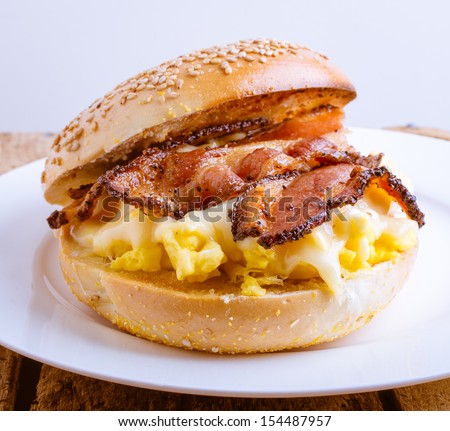Close up of a breakfast sandwich of toasted sesame bagel, slab bacon, egg and melted cheese.