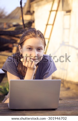 smiling teen girl with laptop sitting at the garden beside of rural house at the evening time