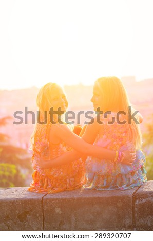 Two twin sisters sitting on the parapet and laugh at sunset. Family composition