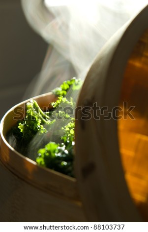 Green leafy vegetables in bamboo steamer with steam in kitchen with moody atmospheric lighting