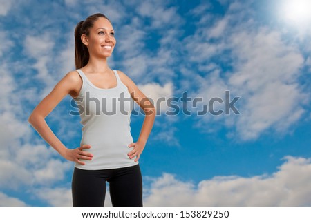 Sporty woman standing up with arms resting at her waist, with a cloudy sky behind her