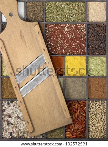 lots of various different spices in a framed dark wooden box with a old wooden mandolin slicer on it, seen from above
