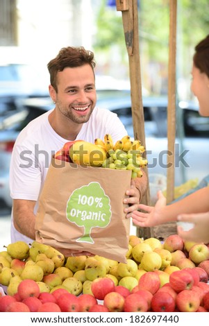 Greengrocer owner of a small business selling fruits and vegetables to a woman carrying a shopping paper bag with a 100% organic certified label.