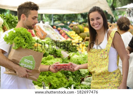 Happy couple shopping fruits and vegetables at a open street market, carrying a shopping paper bag with a 100% organic certified label full of fruit and vegetables.