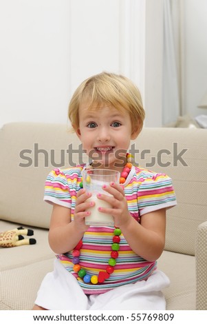 happy smiling cute blond child drink a glass of milk sitting sofa in the living room