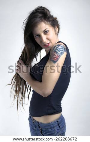 A young female with arm tattoo holding her long hair in white wall/ portrait of girl with tatto in white wall / tattoo of a rose and skull in her arm