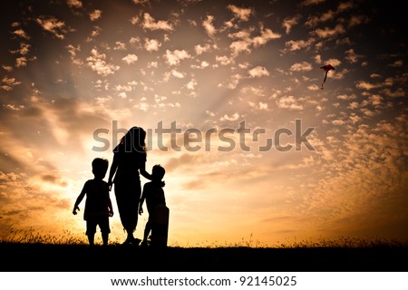 In silhouette, a woman with two young children flying a kite in the sky with a colorful sunset as a backdrop.