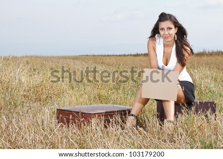A smiling beautiful young woman with long brown hair,wearing a white top and black mini skirt,is sitting in the medow, holding a blank sheet in her hand, her suitcase is next to her, h