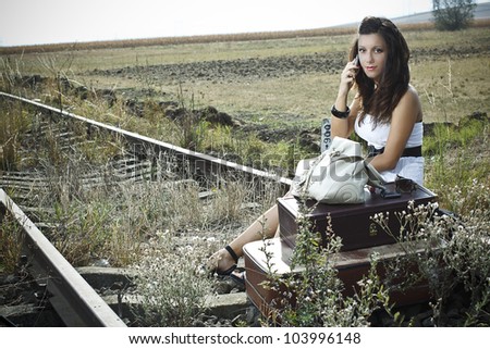 An attractive young woman with long brown hair is sitting on the rail road with old-fashioned suitcases in the front,she is waiting an making a call with her mobile phone