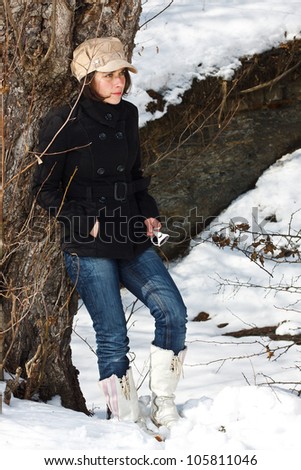 Portrait of a young woman standing in snowy winter forest, wearing a cap, black coat, jeans, white boots, standing by a tree, looking away