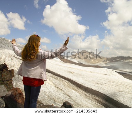 A young girl stands on the edge of a cliff and looking at the sky with clouds and mountain peaks