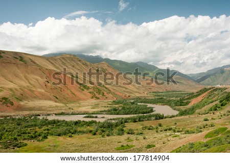 Beautiful river flows in the green valley at the Central Asian mountains covered with white clouds. Bright day in beautiful rural Kyrgyzstan country.