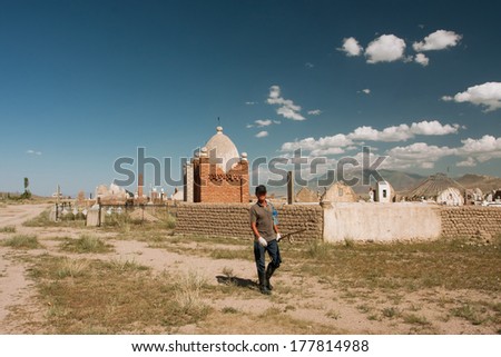KOCHKOR, KYRGYZSTAN - AUG 6: Lonely farmer with scythe goes to a farm on a background of the old Central Asian cemetery on August 6, 2013. Kyrgyzstan is ranked 70th on the Ease of doing Business Index