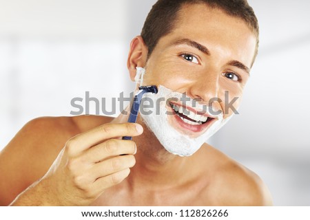 young man shaving in the bath. He is passing the razor for the beard while it looks at the mirror