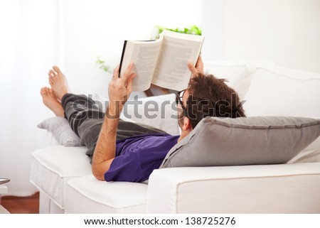 Young man stretching comfortably on couch and reading a book. Man relaxing at home