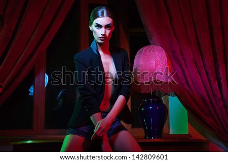 The image of a beautiful fashion woman standing near table with lamp in front of window with curtain