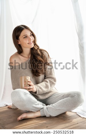 Beautiful girl with cup enjoying the freshness of the new day and looking through the window.