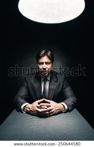 Man in suit sitting in dark room illuminated only by light from a lamp and looking in camera