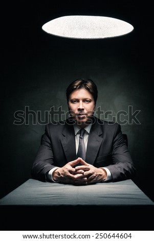 Man in suit sitting in dark room illuminated only by light from a lamp and looking in camera
