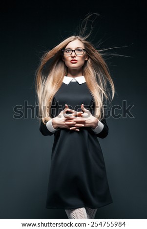portrait of blonde young beautiful posing woman wearing eyeglasses and black dress with white collar. Her fingers crossed.