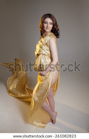 Beautiful young pregnant brunette woman with yellow transparent cloth in studio shot isolated on white background. Model is standing supporting her belly.
