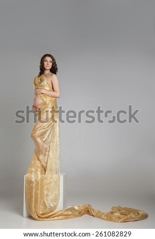 Beautiful young pregnant brunette woman with yellow transparent cloth in studio shot isolated on white background. Model is standing as a statue on cube.