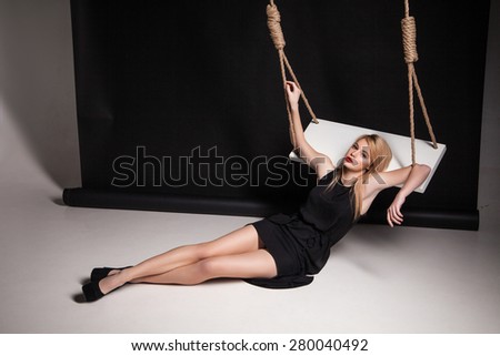 Beautiful young woman in stylish black dress and black shoes half-lying on swing in a studio