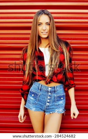 Modern young woman poses in front of red wall background. Sexy woman in jean shorts