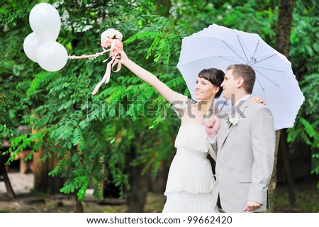 Happy bride and groom standing together in green autumn park holding white balloons in hand