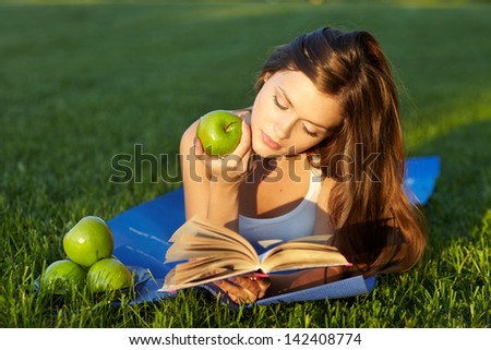 pretty young woman lying on green grass reading book with green apples