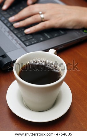 Coffee cup on table with hands and laptop in the background