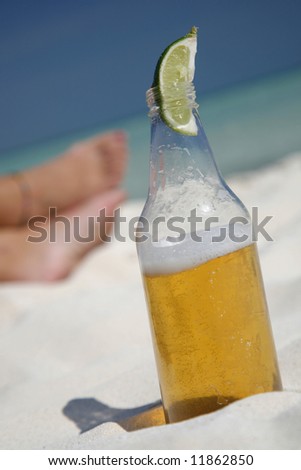 Beer with lime wedge on the beach with women's feet behind