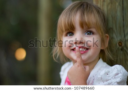 Happy smiling portrait of a 2 year old girl leaning against a wooden post with finger over lips