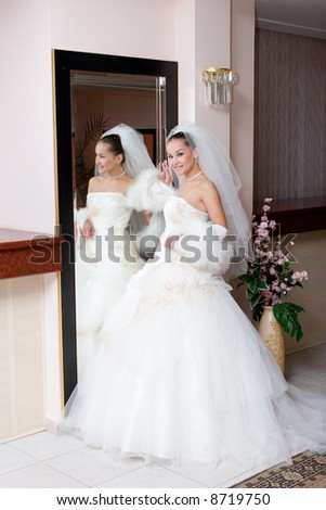 a young smiling bride in a beautiful dress stans by a big mirror