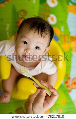 Six month old South East Asian Chinese baby girl sitting in a yellow seat, being spoon fed