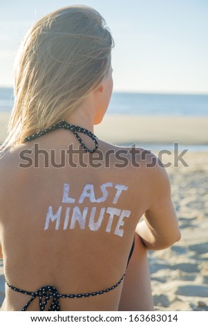 Last Minute written with sunscreen on back of attractive woman sitting on beach looking at ocean