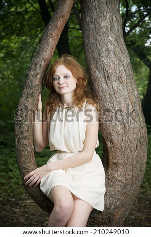 Portrait of redhead girl with blue eyes on nature. Young woman in a dress sitting on a tree